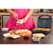 Sandwich maker placed on a tabletop with sandwiches inside nicely toasted. A woman spreading butter on a slice of toast. A wooden board with toast slices on top. And ham, ketchup, cheese slice around the board.